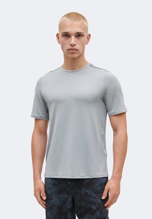 Νέος άνδρας με κοντά ξανθά μαλλιά φοράει ανοιχτό γκρι κοντομάνικο πουκάμισο και σκούρο παντελόνι με σχέδια, στέκεται με το βλέμμα προς τα εμπρός μπροστά σε απλό φόντο.