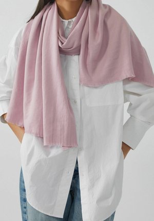 Femme portant une écharpe rose clair drapée sur une chemise blanche à boutons et un jean bleu, debout avec les mains dans les poches.