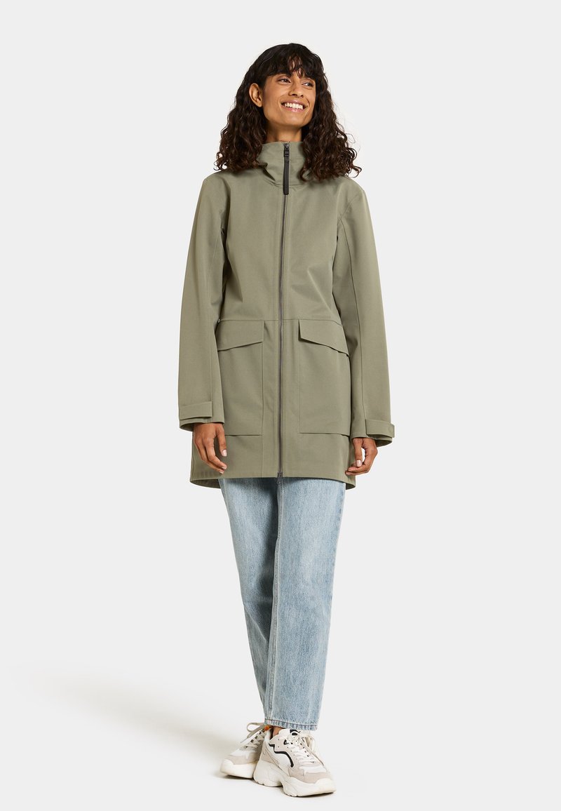 Olivgrüne, wasserdichte Jacke mit hohem Kragen, zwei Fronttaschen und Reißverschluss. Getragen über hellblauen Jeans und weißen Sneakern.