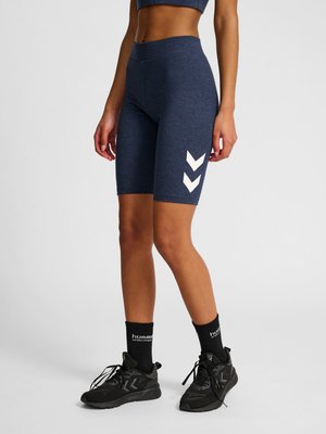 Frau trägt dunkelblaue Bike-Shorts mit hoher Taille und weißen Chevron-Logos, schwarze hummel Sportsocken und schwarze Sportschuhe.