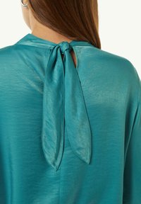 Teal Satinbluse mit einem Bindeband am Nacken und einer kleinen Öffnung. Weiche Textur und lässige Passform, die ein minimalistisches Design unterstreicht.