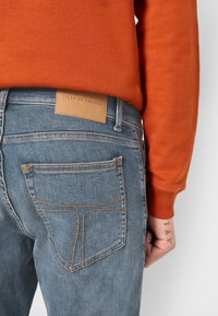 Slim-fit blå denimjeans med orange sömmar och en brun läderetikett. Funktioner inkluderar två bakfickor och en mjuk textur.