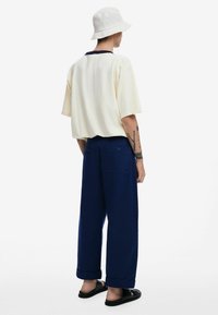 T-shirt oversize beige chiaro, pantaloni a palazzo blu navy, cappello a secchiello bianco e sandali neri. Design semplice senza motivi visibili.