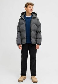 Giacca imbottita grigia con cappuccio, maglione blu navy in maglia, pantaloni neri e scarpe beige. Presenta una chiusura anteriore con cerniera e una texture resistente.