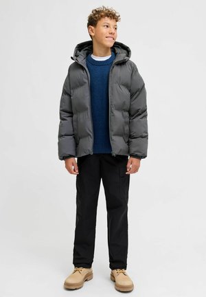 Pelēka pūtēja jaka ar kapuci, tumši zils adīts džemperis, melnas bikses un bēšas kurpes. Aprīkota ar rāvējslēdzēju priekšpusē un izturīgu audumu.