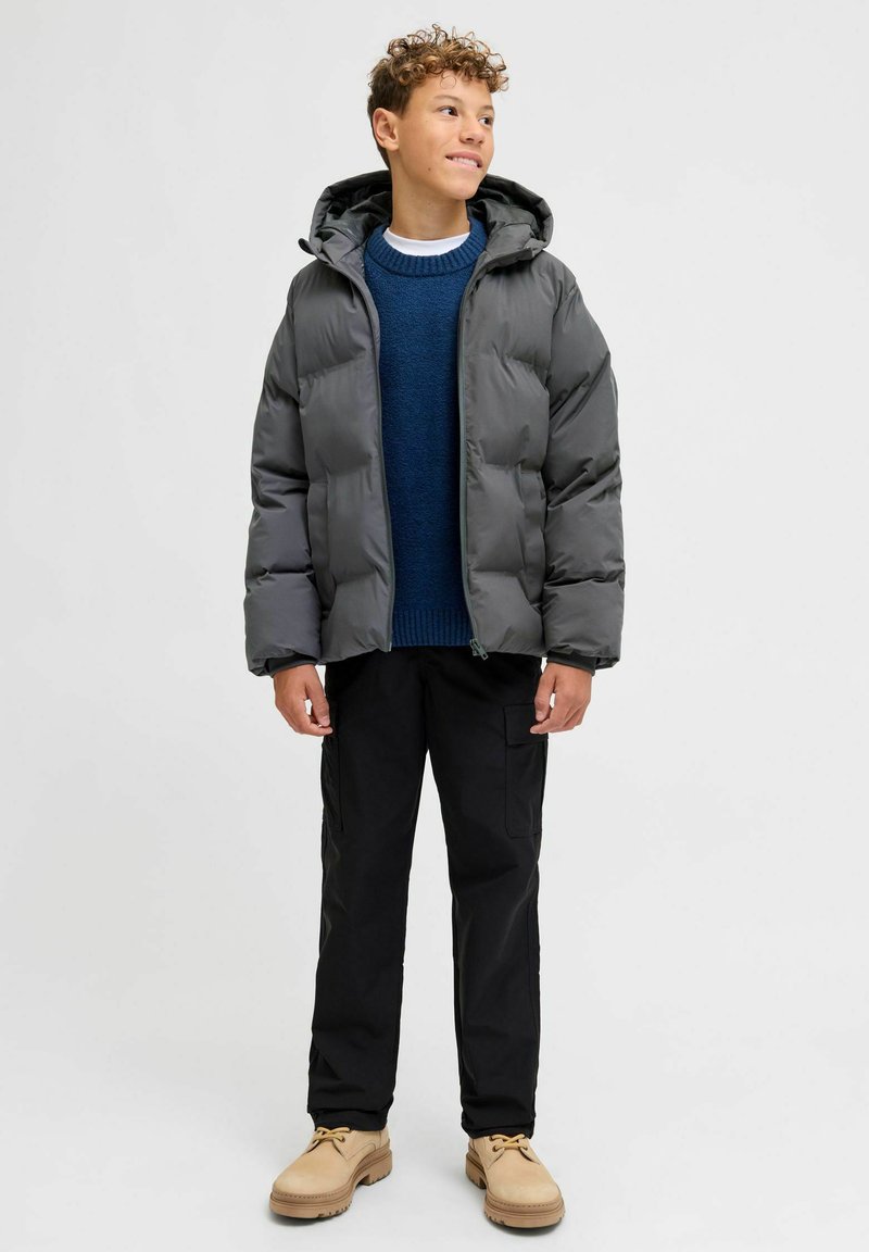 Giacca imbottita grigia con cappuccio, maglione blu navy in maglia, pantaloni neri e scarpe beige. Presenta una chiusura anteriore con cerniera e una texture resistente.