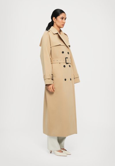 Μπεζ παλτό με διπλή σειρά κουμπιών, μαύρα κουμπιά, ζώνη στη μέση και φαρδιές πατιλέτες, συνδυασμένο με ανοιχτότριχα τζιν και λευκά παπούτσια.