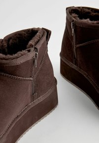 Bottes en daim marron avec un doux doublure en fausse fourrure, présentant des zips sur les côtés et une semelle plateforme, mettant en avant une texture lisse et des lignes épurées.