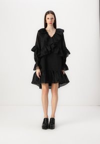 Svart kjole med volanger og lange ermer, laget av teksturert stoff med en utvidet skjortekant. Glinsende svarte platåskjørt fullfører looken.