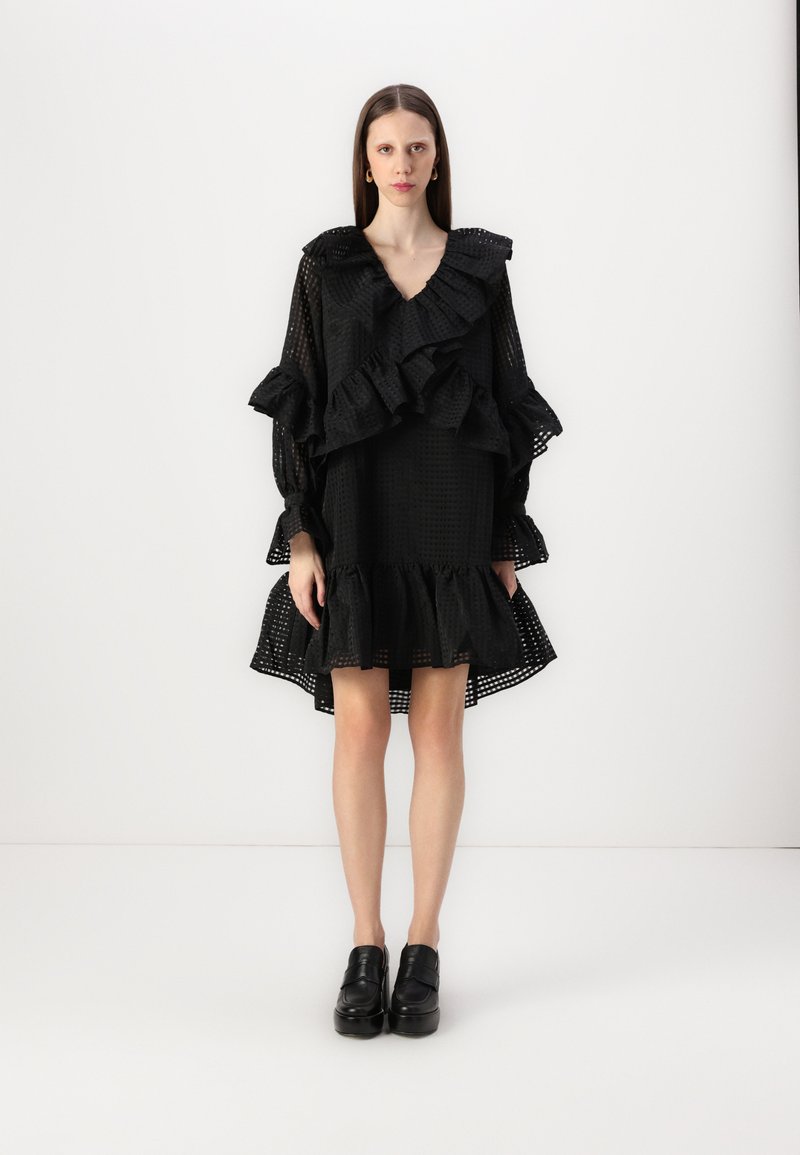 Svart kjole med volanger og lange ermer, laget av teksturert stoff med en utvidet skjortekant. Glinsende svarte platåskjørt fullfører looken.