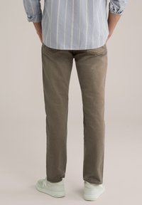 Muškarac u svijetloplavoj prugastoj košulji, smešnim hlačama i bijelim tenisicama, stoji okrenut leđima na bež pozadini.