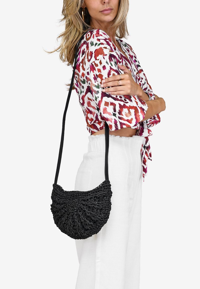 Bolso de mano negro tejido con un diseño curvado y texturizado, y una larga correa negra, llevado al hombro, junto a una blusa con estampado.
