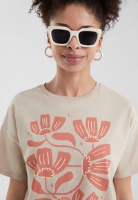 Beige t-shirt featuring a coral floral print design. Model wears white oversized sunglasses and circular earrings. Smooth cotton texture.