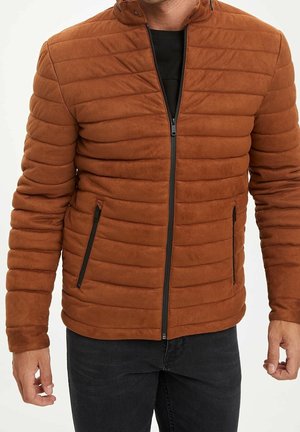 Mann trägt eine rostfarbene, gesteppte Daunenjacke mit schwarzem Reißverschluss und Reißverschlusstaschen an den Seiten, kombiniert mit einer schwarzen Hose.