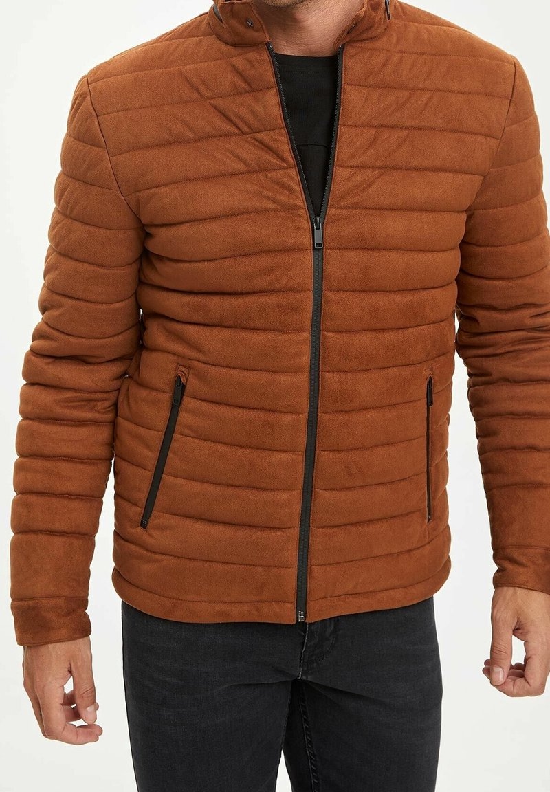 Homme portant une doudoune matelassée couleur rouille avec une fermeture éclair noire et des poches latérales zippées, associée à un pantalon noir.