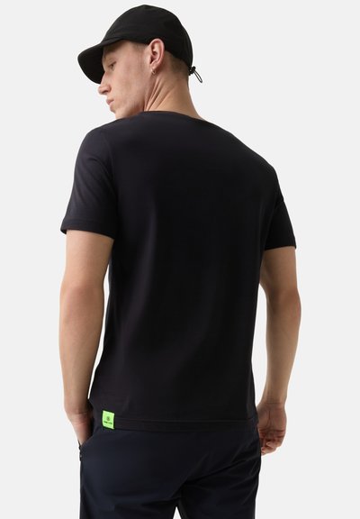 Sort crew-neck t-shirt lavet af glat bomuld. Har et lille grønt mærke ved kanten. Modellen bærer det med mørke bukser og en sort kasket.
