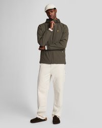 Olivgrüne wasserdichte Jacke mit Kapuze, Reißverschluss und elastischen Bündchen. Kombiniert mit cremefarbenen Cargo-Hosen und dunkelbraunen Slippers.