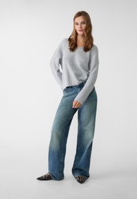 Hellgrauer Strickpullover mit lockerer Passform, kombiniert mit blauen Denim-Jeans im weit geschnittenen Stil. Schwarze Spitzenschuhe mit silbernen Nieten.