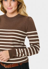 Donna che indossa un maglione a righe marroni e crema con spalle a sbuffo e una cintura marrone, su uno sfondo bianco.