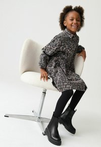 Mlada djevojka s kovrčavom kosom sjedi na modernoj bijeloj stolici, nosi haljinu s leopardovim uzorkom, crne hulahopke i crne čizme, smiješi se u kameru.