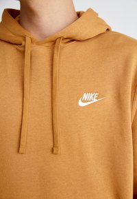 Nike džemperis siltā, sinepju krāsā. Mīksts audums ar kangaroo kabatu, kapuci ar aizdari un balu izšuvumu uz krūtīm.