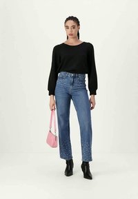 Jeans in denim blu con applicazioni di perle all'orlo, abbinati a un top nero e una borsa rosa. La modella indossa stivali neri alla caviglia.