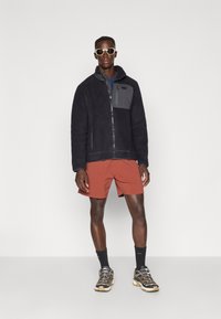Schwarze Fleecejacke mit einem grauen Patch, Reißverschluss, kurze rote Shorts und schwarze sowie beige Wanderschuhe. Die Accessoires umfassen weiße Sonnenbrillen.