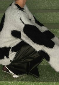 Osoba ubrana w czarny i biały futrzany płaszcz w wzory, czarne spodnie i srebrne szpilki siedzi na zielonych, strukturalnych schodach.