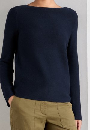 Persoon draagt een marineblauwe trui met lange mouwen en olijfgroene broek, staand met handen in de zakken tegen een effen muur.