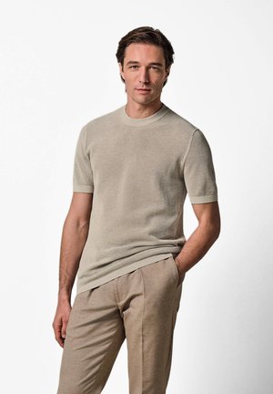 Homme aux cheveux bruns portant une chemise en tricot beige à manches courtes et un pantalon beige assorti, se tenant devant un fond blanc.