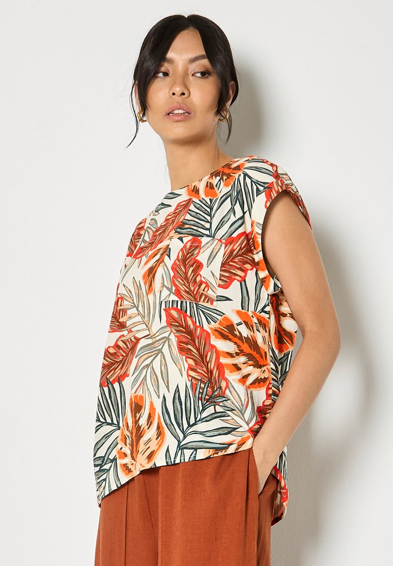 Femme portant un haut sans manches à imprimé feuilles tropicales et un pantalon couleur rouille, la main dans la poche, regardant sur le côté.