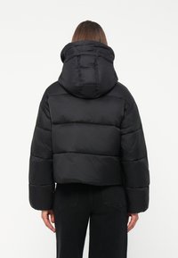 Zwarte pufferjack met een cropped snit, heeft een grote capuchon, een gewatteerd patroon en oversized mouwen. Zachte, gladde textuur.