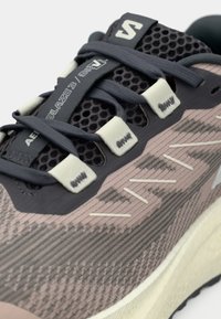 Κοντινό πλάνο αθλητικού παπουτσιού που δείχνει σκοτεινά γκρίζα κορδόνια, γλώσσα από δίχτυ και υφασμάτινο επάνω μέρος με υφή, με το γράμμα "AERO BLAZE 3 GRVL" τυπωμένο στη γλώσσα.