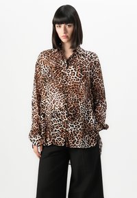 Mulher com cabelo preto liso veste uma camisa de botão solta com padrão de leopardo e calças largas pretas contra um fundo branco liso.