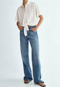 Hellbeige Kurzarmhemd, das an der Taille gebunden ist, kombiniert mit blauen weit geschnittenen Jeans. Der Stoff wirkt weich und hat eine glatte Textur.