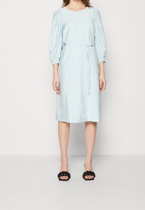 Robe midi bleu clair avec manches trois-quarts bouffantes, ceinture nouée à la taille et sandales noires tressées à talons ouverts.