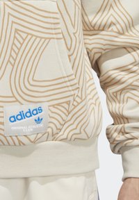 Beige mikina Adidas s abstraktními vzory světle hnědé barvy, klokankou a bílou etiketou s nápisem "adidas Original Athletic Club" v modré a bílé.