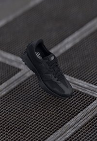 Crni New Balance tenisice suspendirane iznad metalne mrežaste površine s čeličnim poprečnim trakama u industrijskom okruženju.