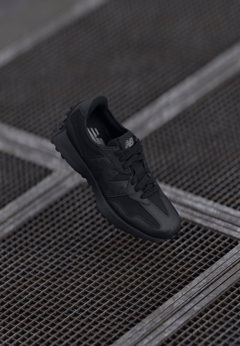Svart New Balance sneakers svevende over en metallgitteroverflate med stål tverrbjelker i et industrielt miljø.