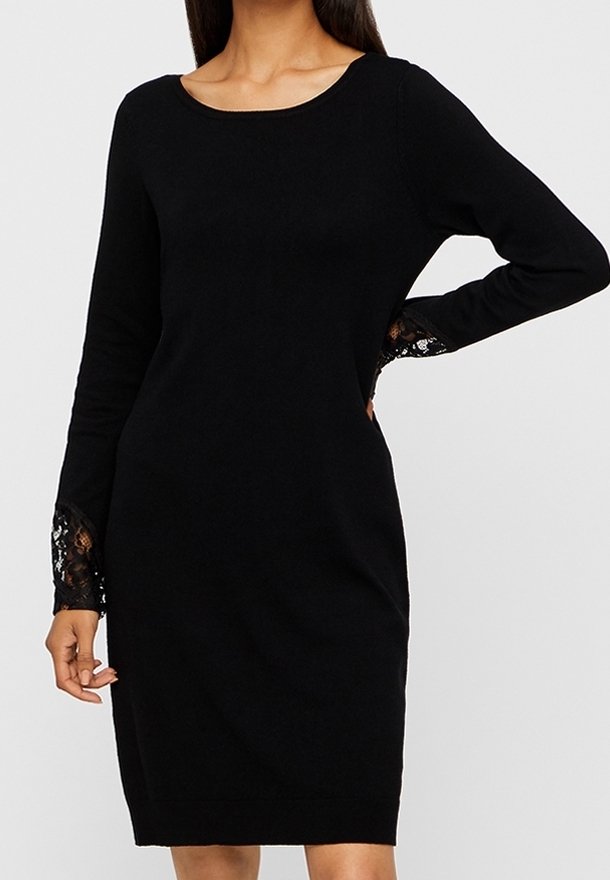 Femme portant une robe noire cintrée à manches longues avec des détails en dentelle sur les poignets, debout avec une main sur la hanche.