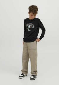 Felpa nera con grafiche "ORIGINALS" e design di teschio, abbinata a pantaloni beige a gamba larga e sneakers bianco e nere.