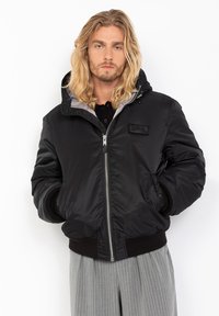 Zwarte nylon bomberjack met een capuchon, voorzien van een zilveren rits, geribbelde manchetten en een logo patch op de linkerkant van de borst.