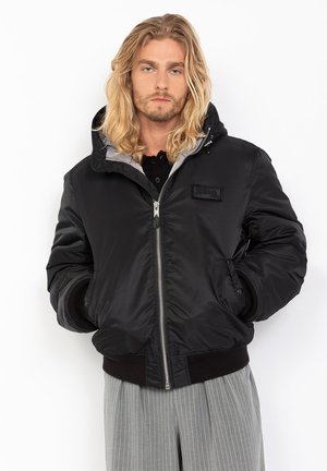 Zwarte nylon bomberjack met een capuchon, voorzien van een zilveren rits, geribbelde manchetten en een logo patch op de linkerkant van de borst.