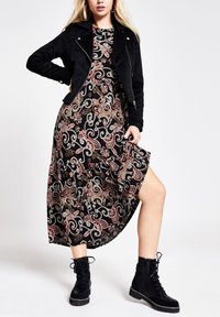 Donna che indossa una giacca biker in suede nera sopra un vestito midi a fantasia nera, sollevando il vestito per mostrare stivali neri alla caviglia con lacci e suole spesse.