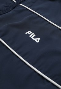Laivastonsininen FILA-takin yksityiskohta, jossa valkoinen nauhakuosi ja vetoketju, ja valkoinen FILA-logo keskellä kangasta.