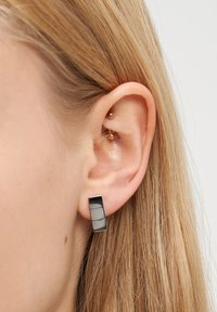 Nahaufnahme eines linken Ohrs mit blonden Haaren, das ein kleines goldenes Double-Ball-Cartilage-Piercing und einen rechteckigen schwarzen und silbernen Ohrring zeigt.