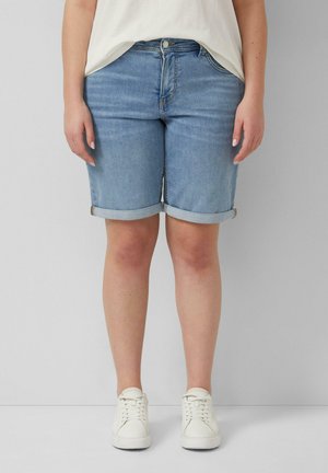 Person steht und trägt hellblaue Jeansshorts mit umgeschlagenen Säumen, weiße Sneakers und ein weißes Top vor einem einfarbigen grauen Hintergrund.