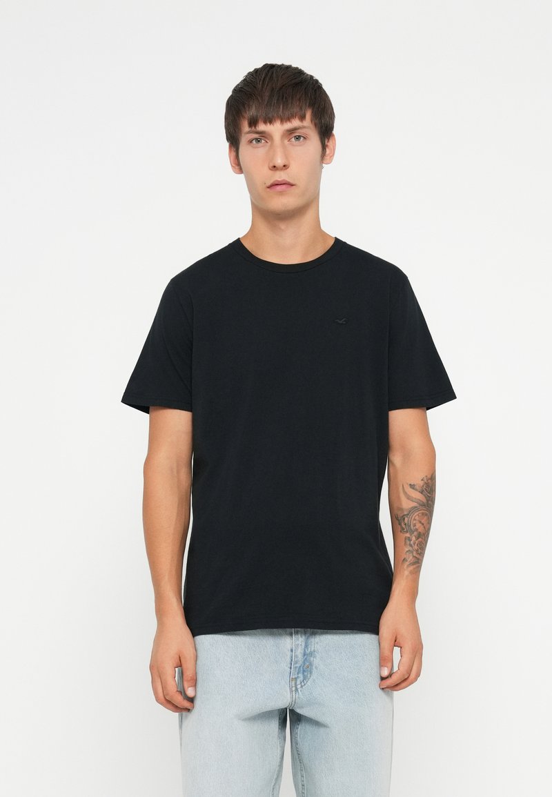 T-shirt nera in cotone con scollatura rotonda e maniche corte. Presenta un logo minimalista ricamato sul petto. Abbinata a jeans azzurri chiari.