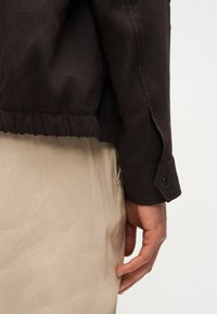 Άτομο που φοράει σκούρο μακρυμάνικο πουκάμισο με κουμπωμένο μανίκι και ανοιχτόχρωμο μπεζ παντελόνι, με το χέρι να ακουμπάει δίπλα στο σώμα του σε απλό φόντο.