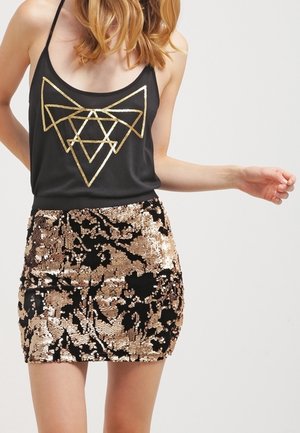 Kobieta w czarnej, bezrękawnikowej bluzce z geometrycznym, złotym wzorem i czarnej mini spódnicy pokrytej złotymi cekinami na jednolitym tle.